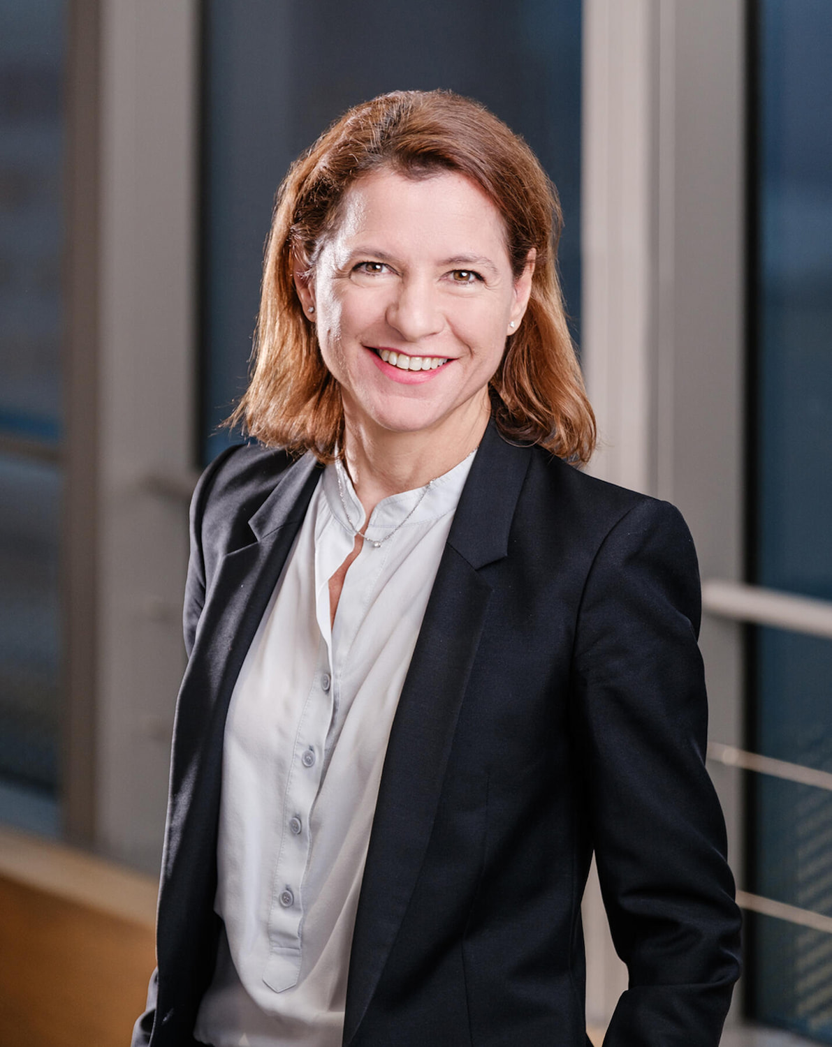 Professional woman in a dark suit and light blouse, standing indoors with a modern background.